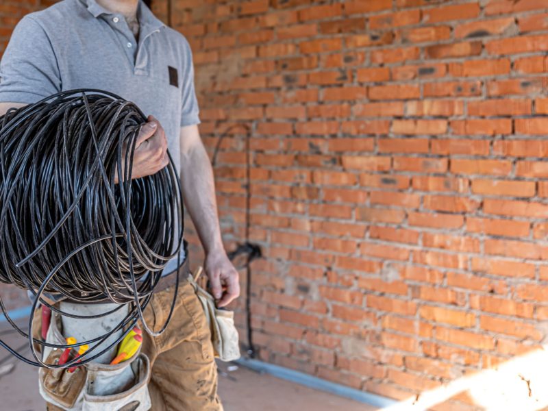 An electrician in a hard hat looks at the wall while holding an electric cable.