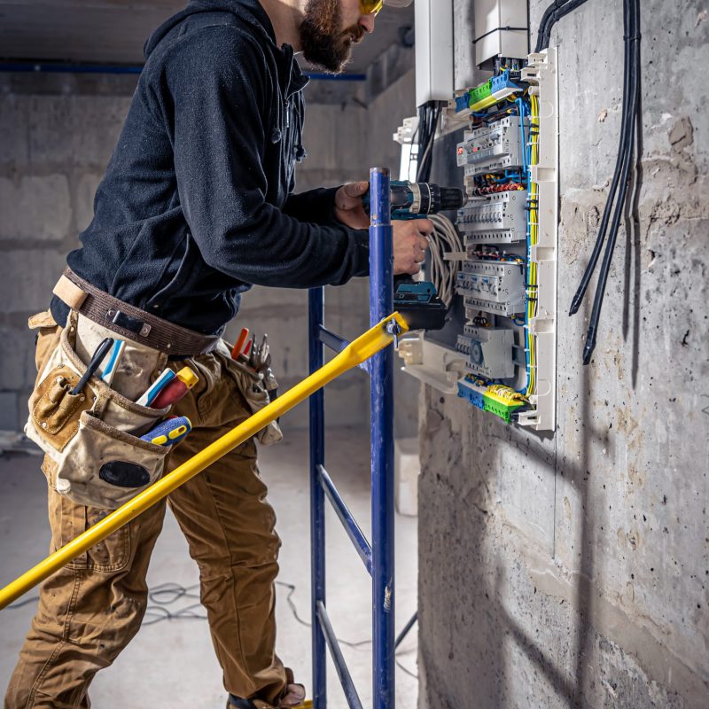 A male electrician works in a switchboard with an electrical connecting cable, connects the equipment with tools.